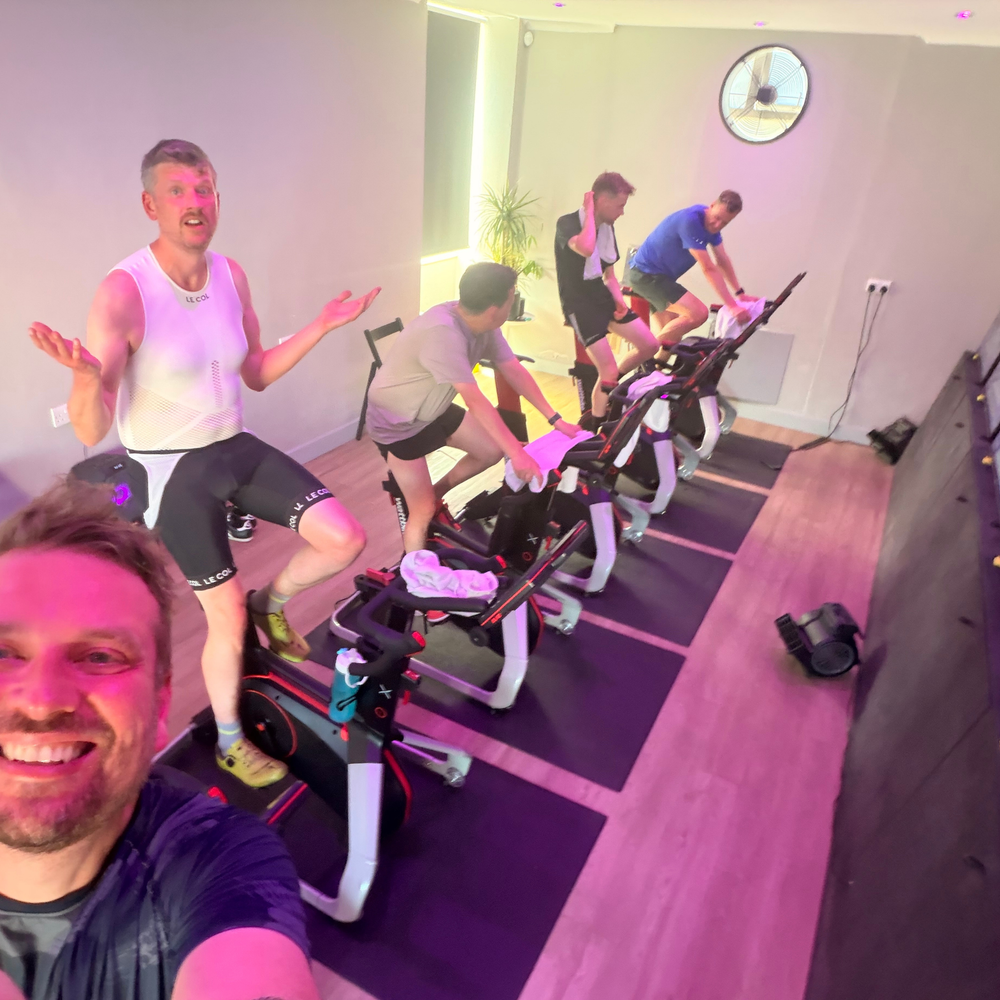 A group of people riding indoor stationary bikes in a fitness studio with pink ambient lighting. One man in the foreground is taking a selfie, while others exercise on black and white bikes arranged diagonally across the room.