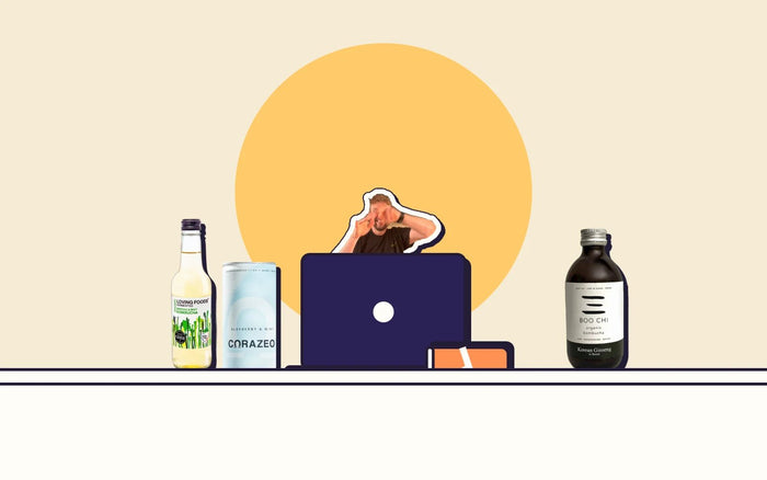 A desk with a laptop and three healthy drinks, a sugary energy drink knocked over, and Kombucha Kev peeking over the screen