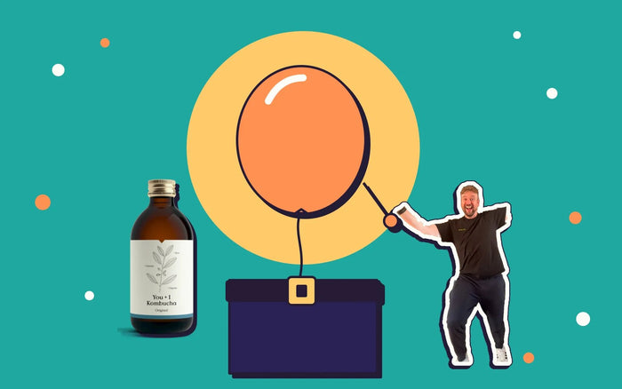 A balloon tied to a belt, a bottle of You + I kombucha, and Kombucha Kev creeping up with a pin