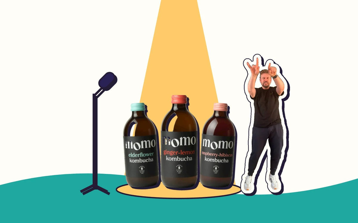 Three bottles of MOMO kombucha under a stage spotlight with a mic stand and Kombucha Kev rocking out