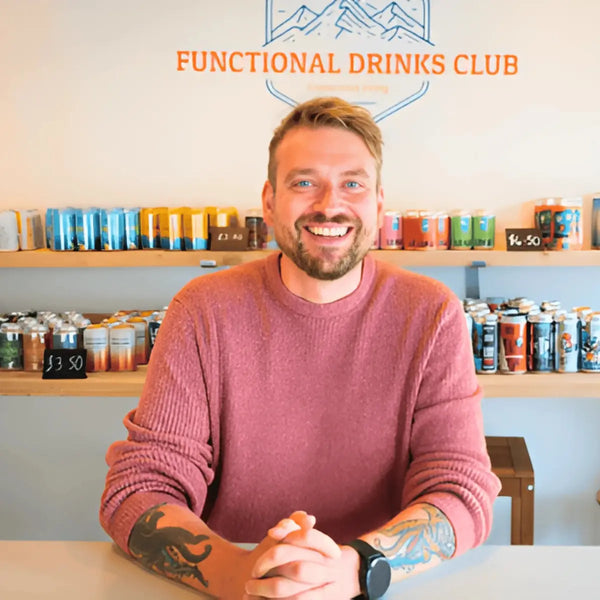 Kombucha Kev, founder of Functional Drinks Club, smiling at the counter of the Otley taproom in a pink jumper, with shelves of drinks and the Functional Drinks Club sign behind him.