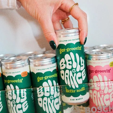 A hand picking a can of Belly Dance Kiwi Mango water kefir from a fridge stacked with cans.