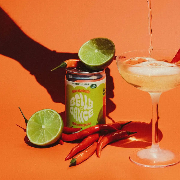 A can of Belly Dance Spicy Margarita water kefir beside a coupe glass, with fresh limes and red chillies on an orange background.