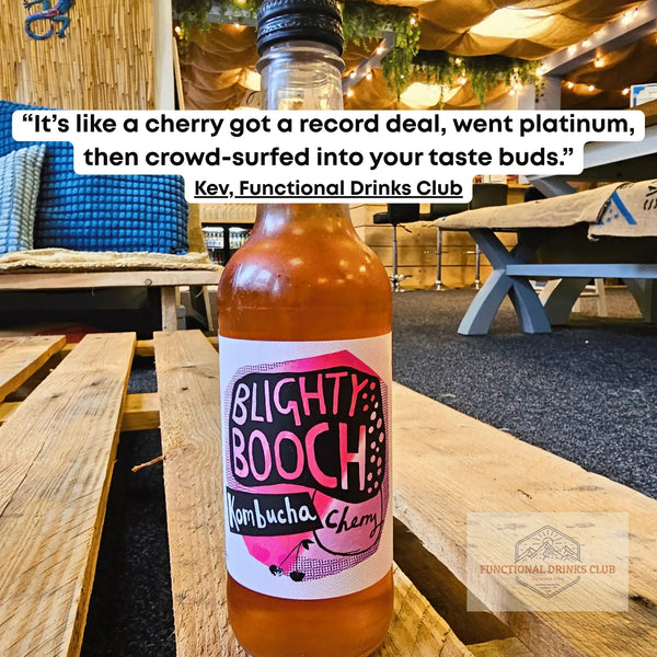 Blighty Booch Cherry raw kombucha on a wooden pallet table in the Otley taproom, with Kev's tasting note: "It's like a cherry got a record deal, went platinum, then crowd-surfed into your taste buds."