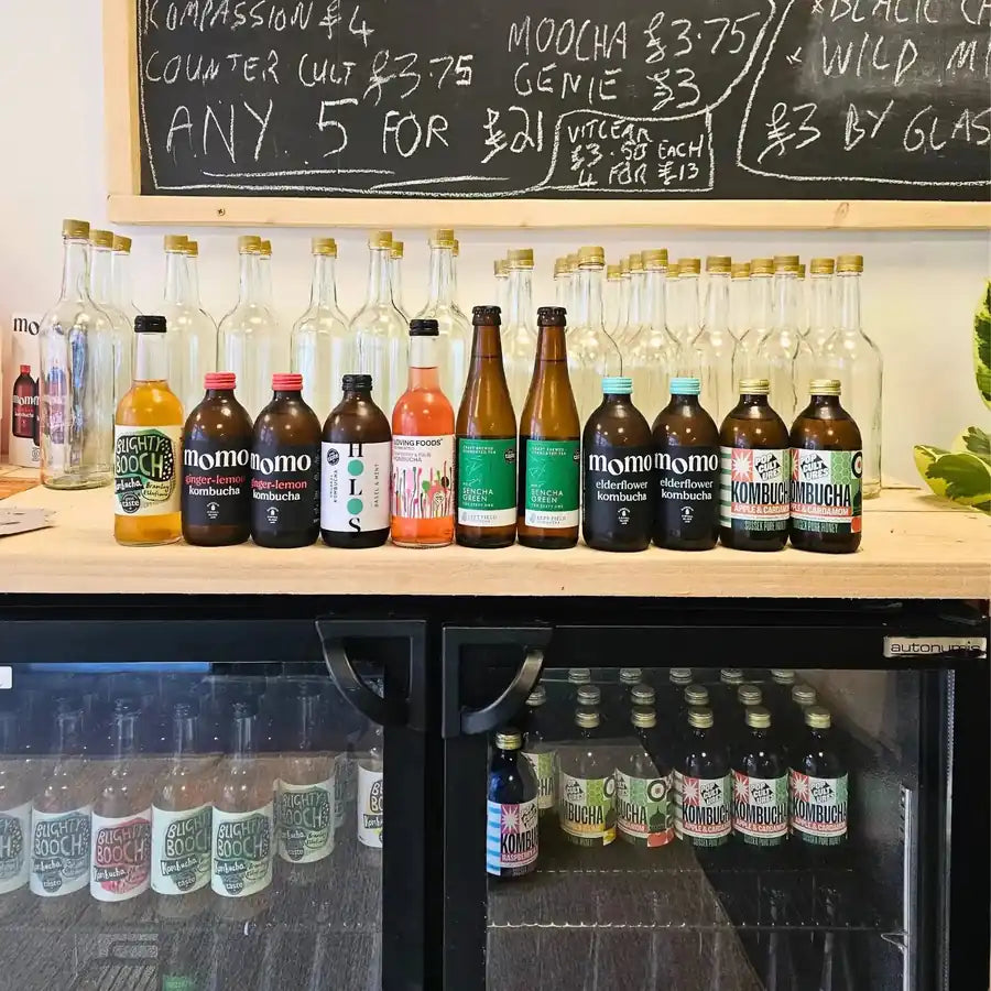 Raw kombucha from the makers we stock lined up on the counter above the drinks fridge in the Otley taproom, with the chalkboard price list behind.