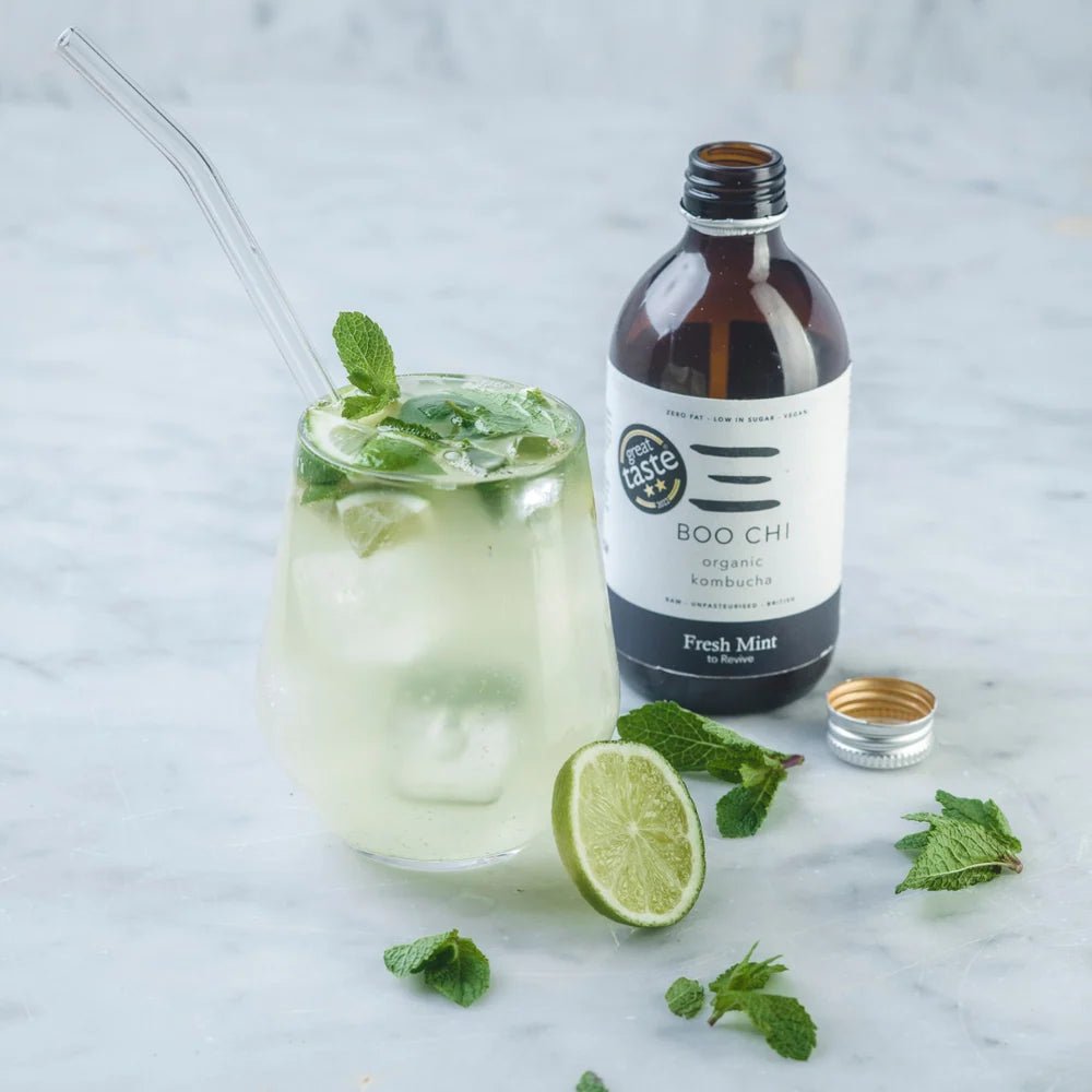 A bottle of Boo Chi Fresh Mint raw kombucha beside a glass with ice, fresh mint, lime and a glass straw on white marble.