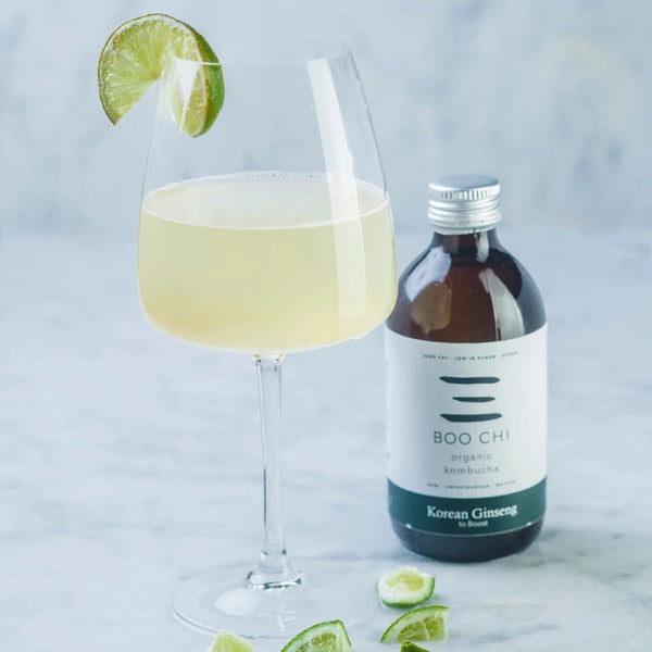 A bottle of Boo Chi Korean Ginseng raw kombucha beside a wine glass of kombucha with a lime wedge on white marble.