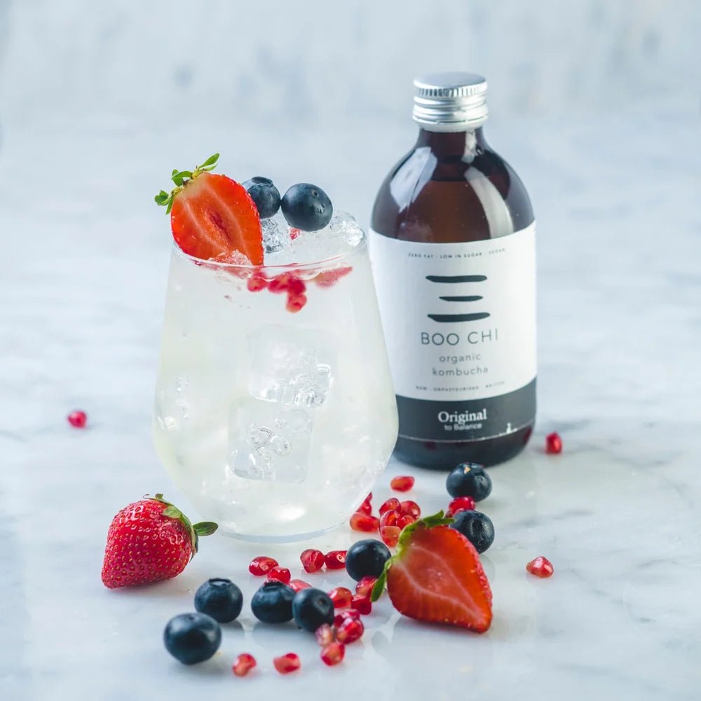 A bottle of Boo Chi Original raw kombucha beside a glass of kombucha over ice, with strawberries, blueberries and pomegranate seeds on white marble.