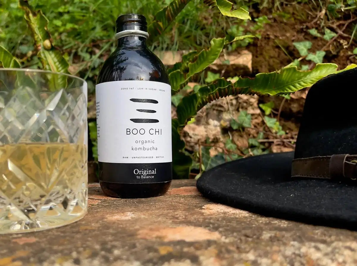 A bottle of Boo Chi Original raw kombucha outdoors on a stone ledge among leaves, beside a cut-glass tumbler and a black hat.