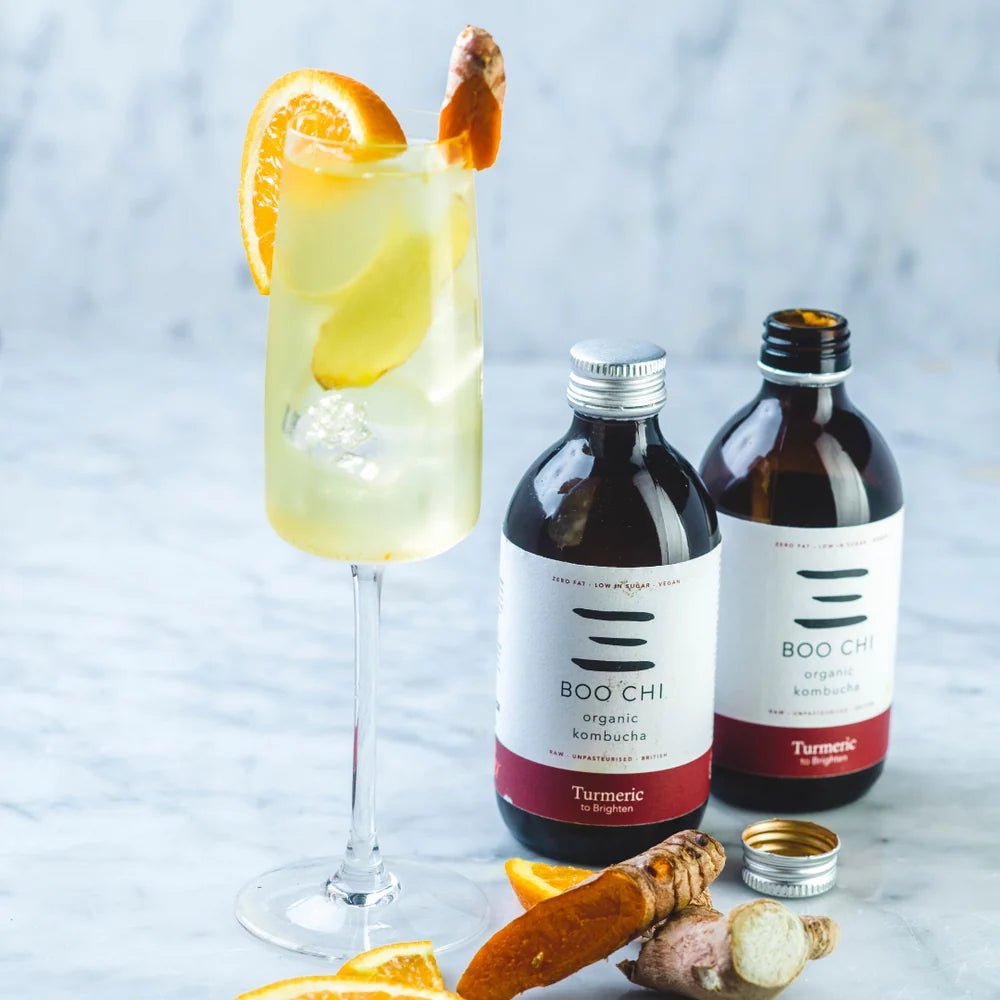 Two bottles of Boo Chi Turmeric & Black Pepper raw kombucha beside a tall glass with an orange slice, with fresh turmeric root on white marble.