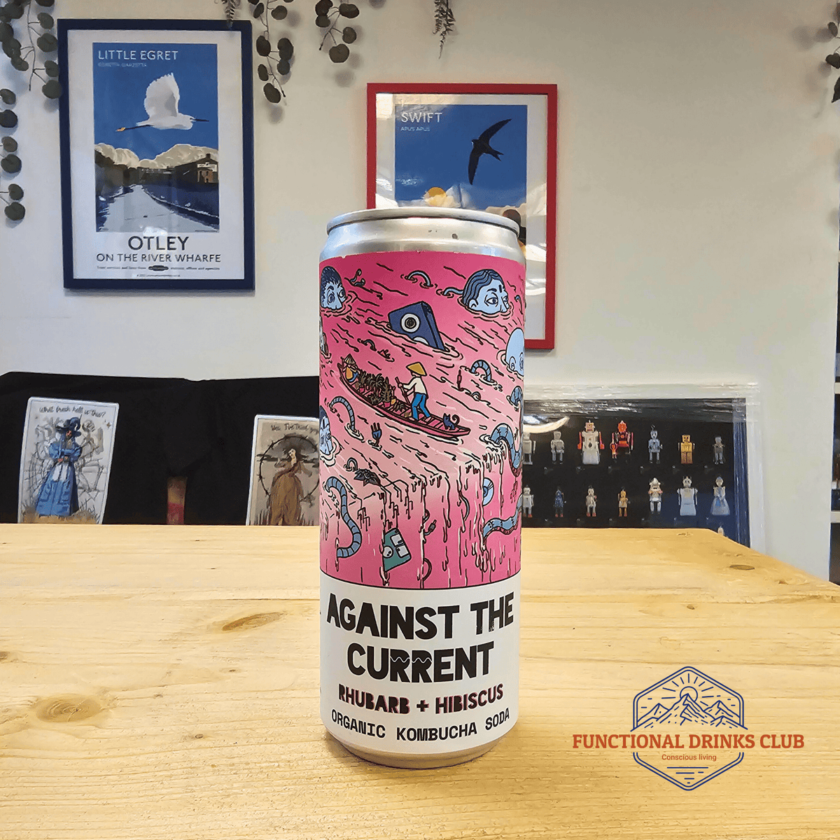 A can of Counter Culture Rhubarb & Hibiscus raw kombucha on a wooden table in front of framed Otley art prints.