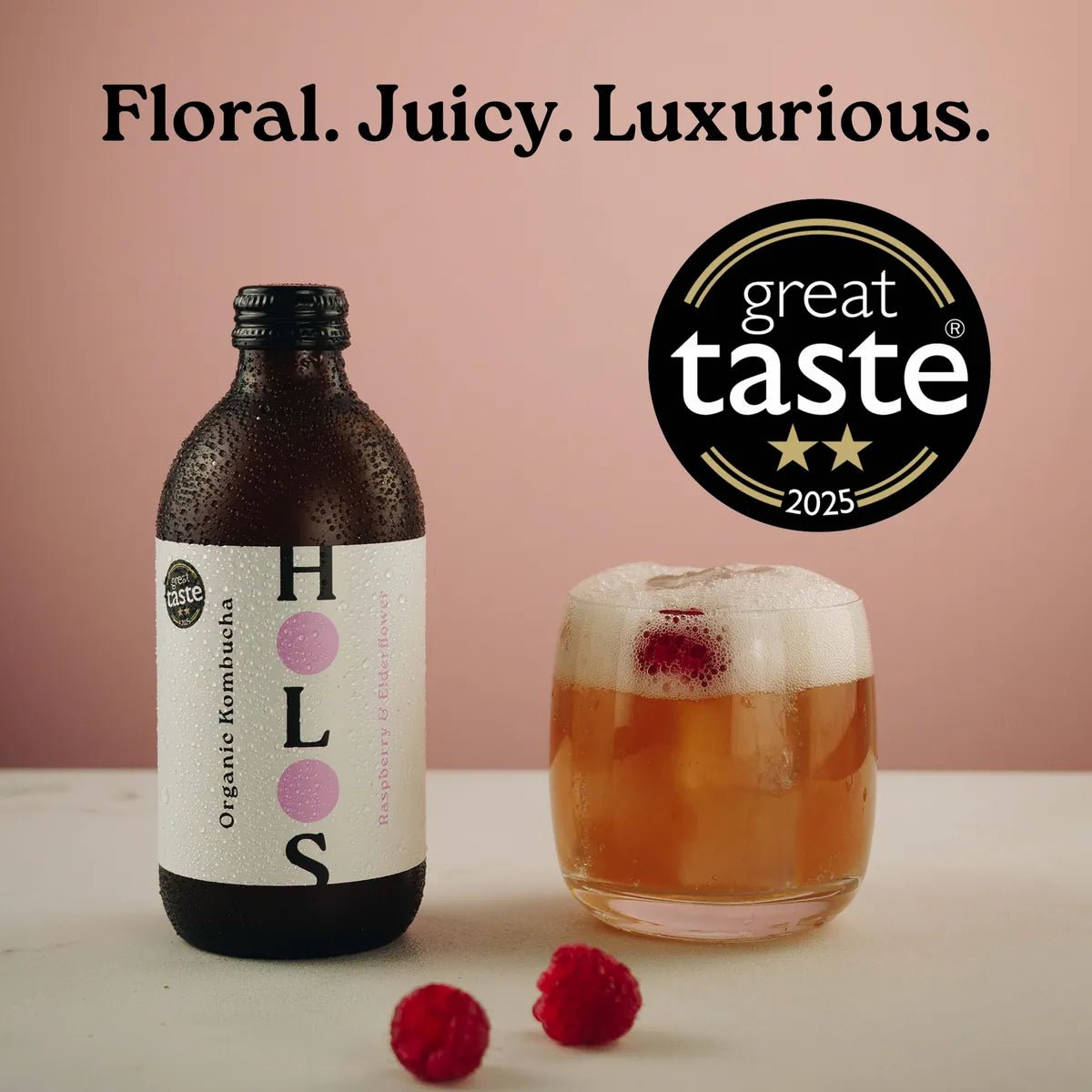 A bottle of HOLOS Raspberry & Elderflower kombucha beside a glass with a raspberry on top, with fresh raspberries and a 2-star Great Taste 2025 badge.
