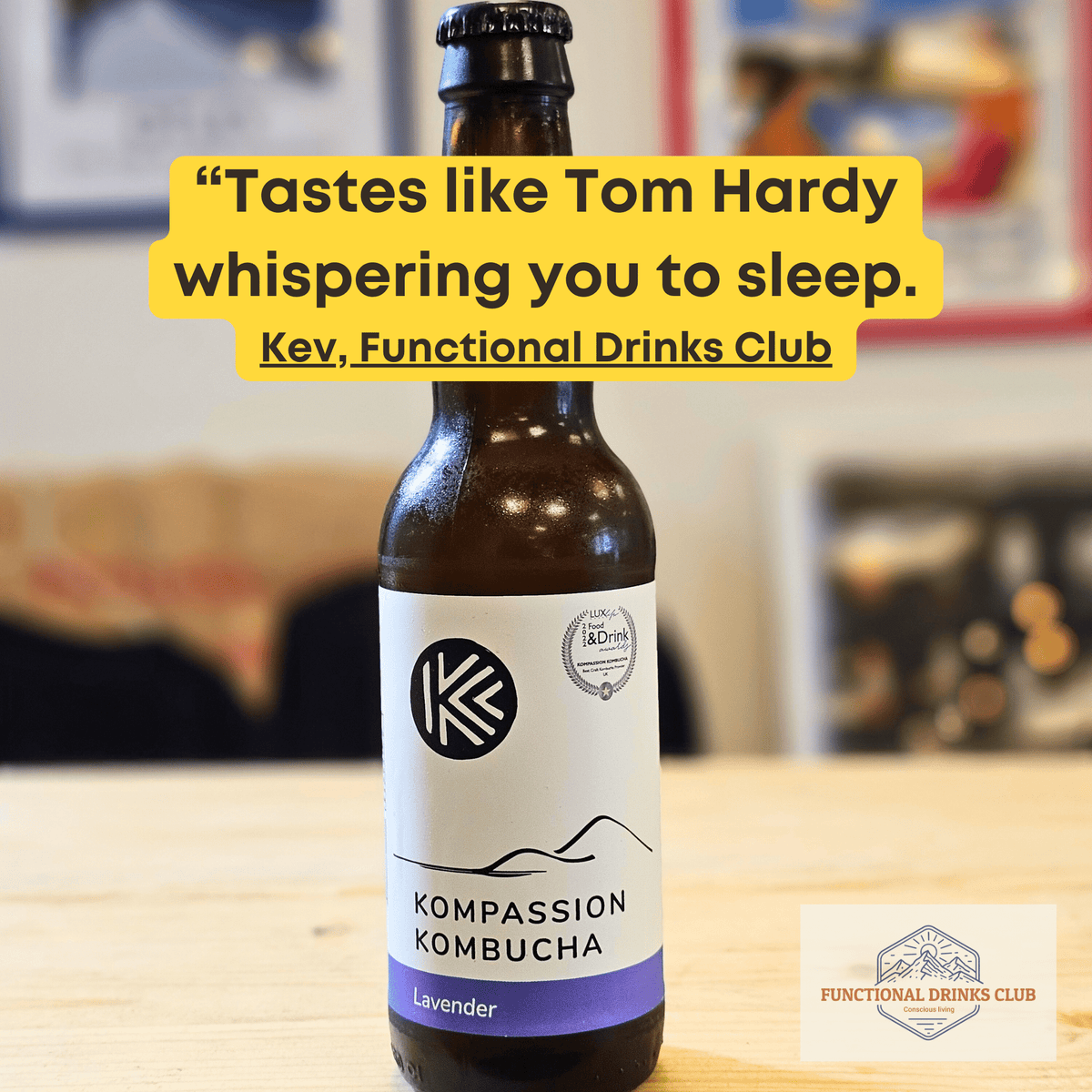 Kompassion Lavender raw kombucha on a wooden table with framed prints softly blurred behind, and Kev's tasting note: "Tastes like Tom Hardy whispering you to sleep."