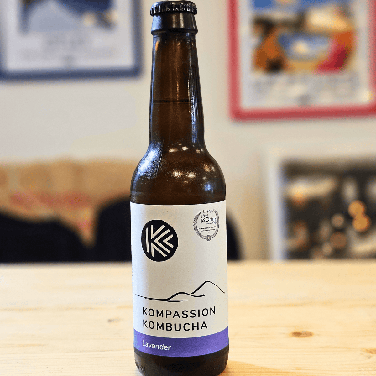 A bottle of Kompassion Lavender raw kombucha on a wooden table with framed prints softly blurred behind.