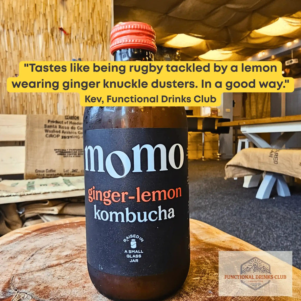 MOMO Ginger & Lemon raw kombucha on a tree-trunk table in the Otley taproom, with Kev's tasting note: "Tastes like being rugby tackled by a lemon wearing ginger knuckle dusters. In a good way."