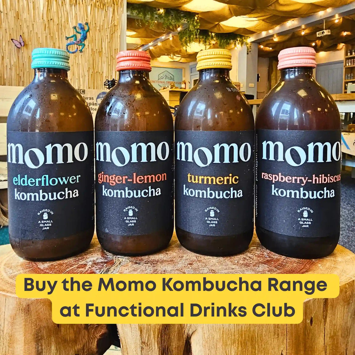 The four MOMO raw kombucha flavours, Elderflower, Ginger & Lemon, Turmeric and Raspberry & Hibiscus, lined up on a tree-trunk table in the Otley taproom under a yellow banner: "Buy the Momo Kombucha Range at Functional Drinks Club".