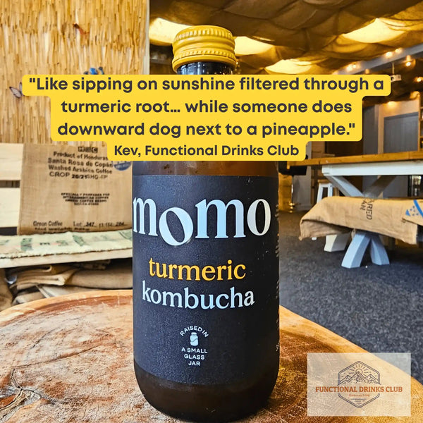 MOMO Turmeric raw kombucha on a tree-trunk table in the Otley taproom, with Kev's tasting note: "Like sipping on sunshine filtered through a turmeric root... while someone does downward dog next to a pineapple."
