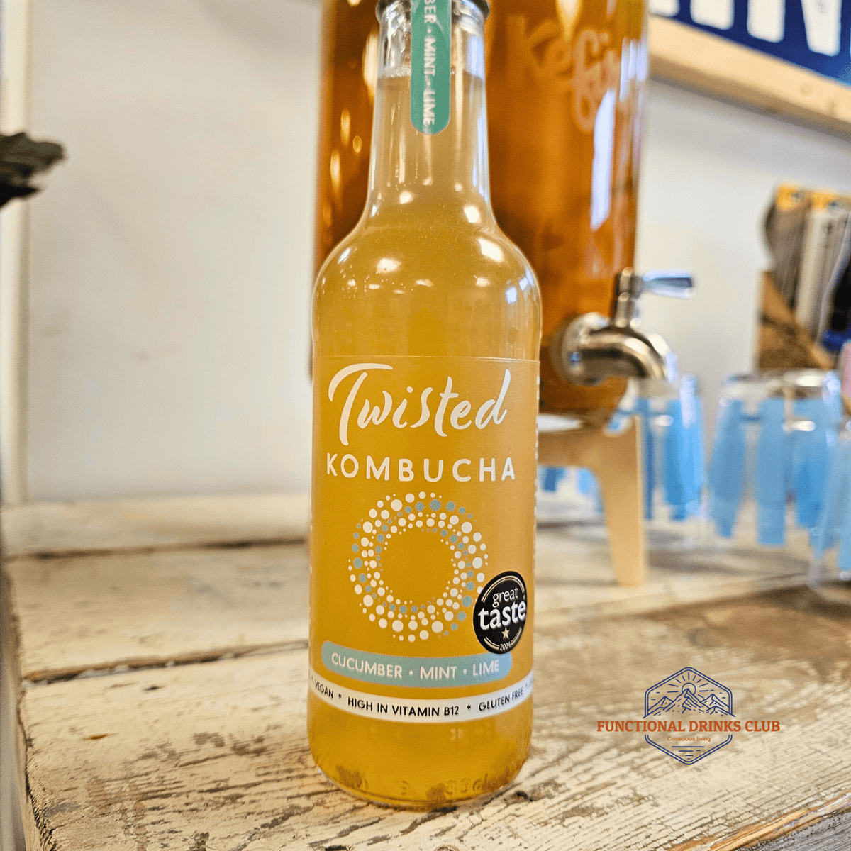 A bottle of Twisted Cucumber, Mint & Lime raw kombucha on a rustic wooden counter in the Otley taproom, with a glass kombucha dispenser behind.