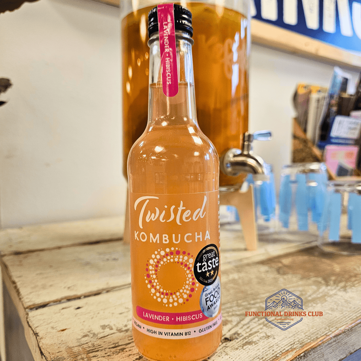 A bottle of Twisted Hibiscus & Lavender raw kombucha on a rustic wooden counter in the Otley taproom, with a glass kombucha dispenser behind.