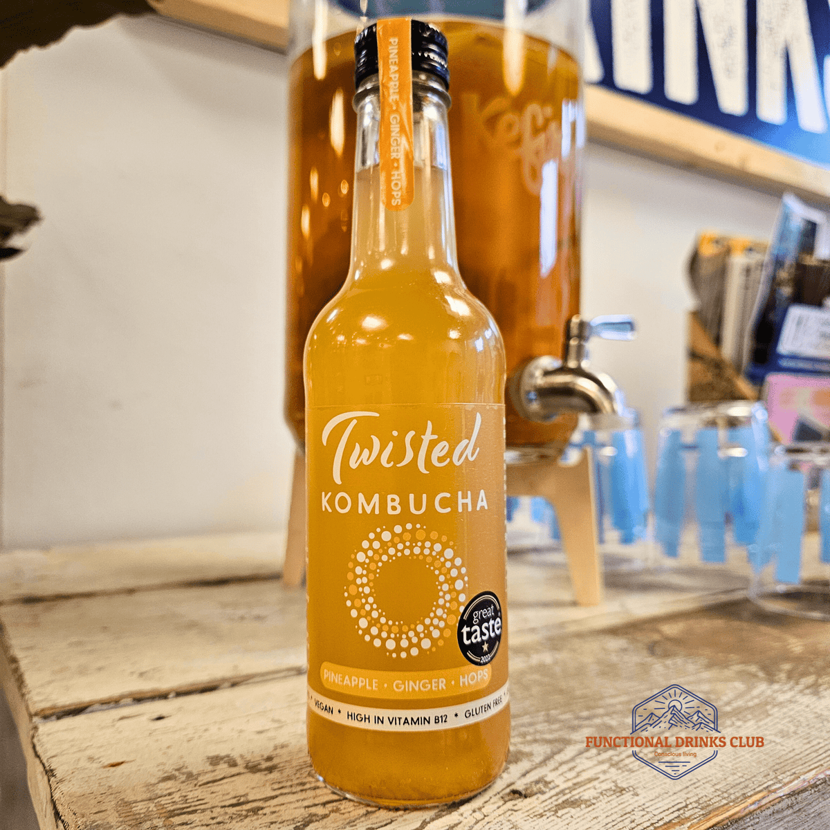 A bottle of Twisted Pineapple, Ginger & Hops raw kombucha on a rustic wooden counter in the Otley taproom, with a glass kombucha dispenser behind.