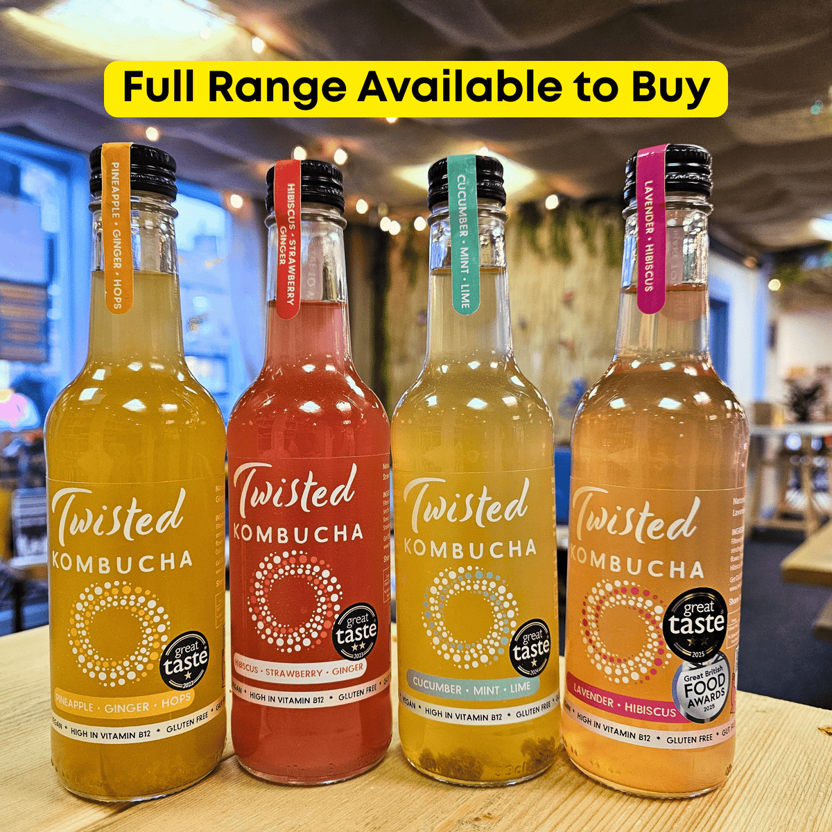 All four Twisted raw kombucha flavours lined up on a wooden table in the Otley taproom under a yellow 'Full Range Available to Buy' banner.