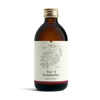 You + I Black Cherry raw kombucha in an amber glass bottle with a gold cap and a cream label with a botanical line drawing and a deep red band.
