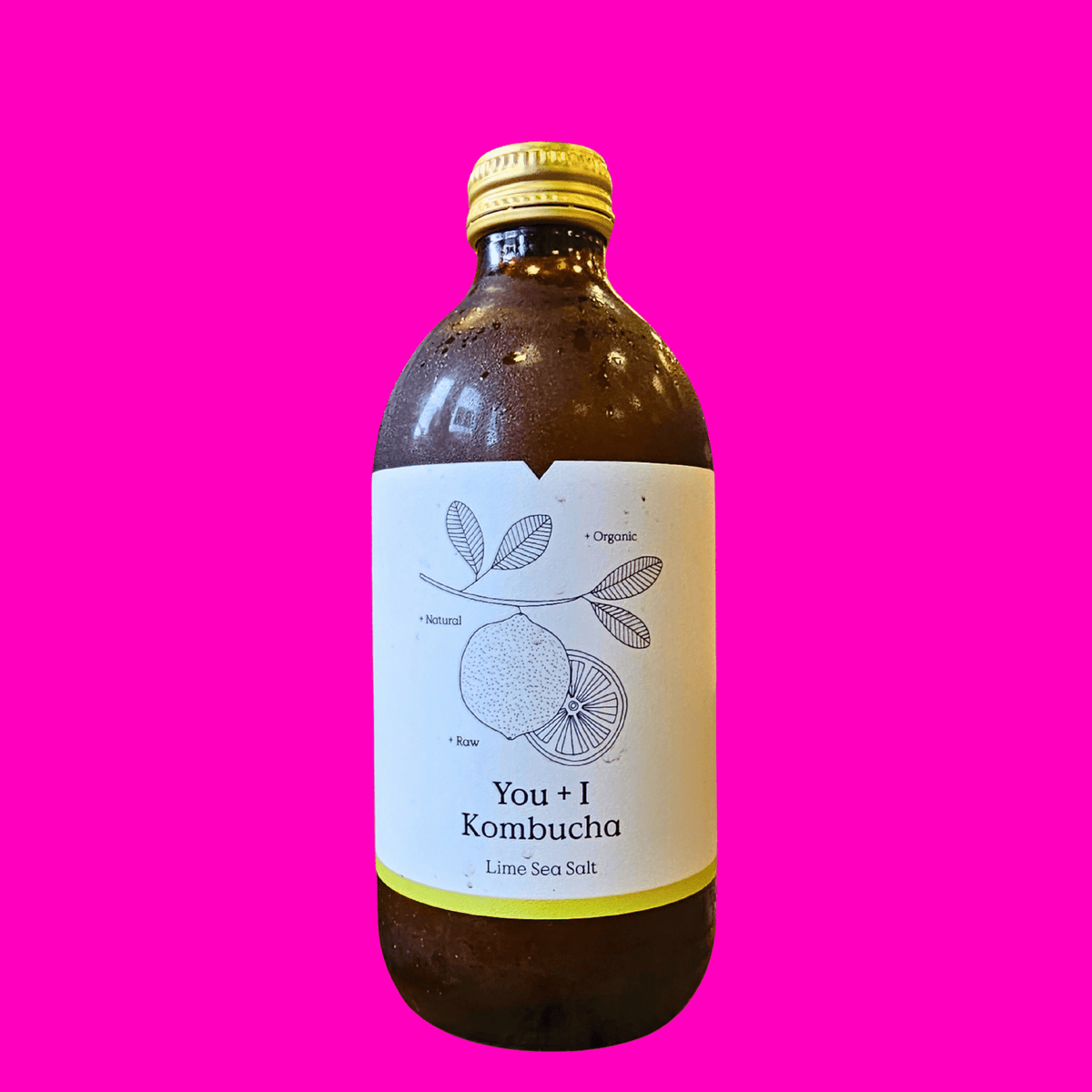 A bottle of You + I Lime & Sea Salt raw kombucha on a hot pink background.